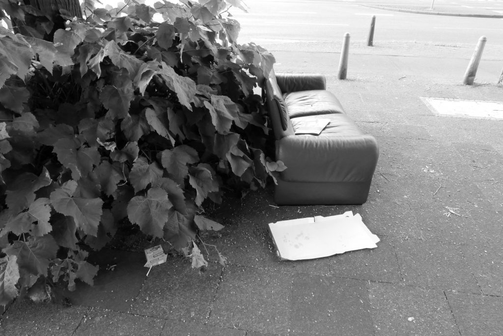 Ein Sofa auf dem Gehweg. Daneben eine leere Tüte ,,Durstlöscher'', sowie eine leere Pizzaschachtel. Auf dem Sofa eine Zeitung.