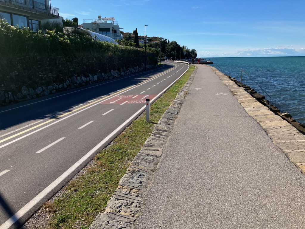 Küstenstraße. Linker Fahrstreifen ist für Autos, der rechte Fahrstreifen ist für Fahrräder (unterteilt für Gegenverkehr). Rechts daneben, ein breiter Fußweg. Daneben das Meer.