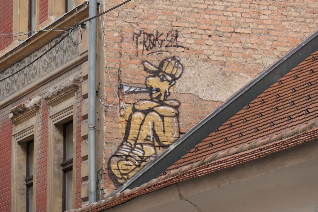 Auf einer Häuserwand ein Graffito eines kiffenden Mannes. Es wirkt so, als würde er auf einer Dachschräge sitzen.