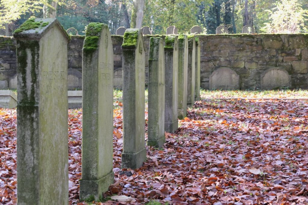 Grabsteine in einer Reihe. Man sieht im Hintergrund eine Mauer, davor weitere Grabsteine. Hinter der Mauer lugen noch mehr Grabsteine hervor.