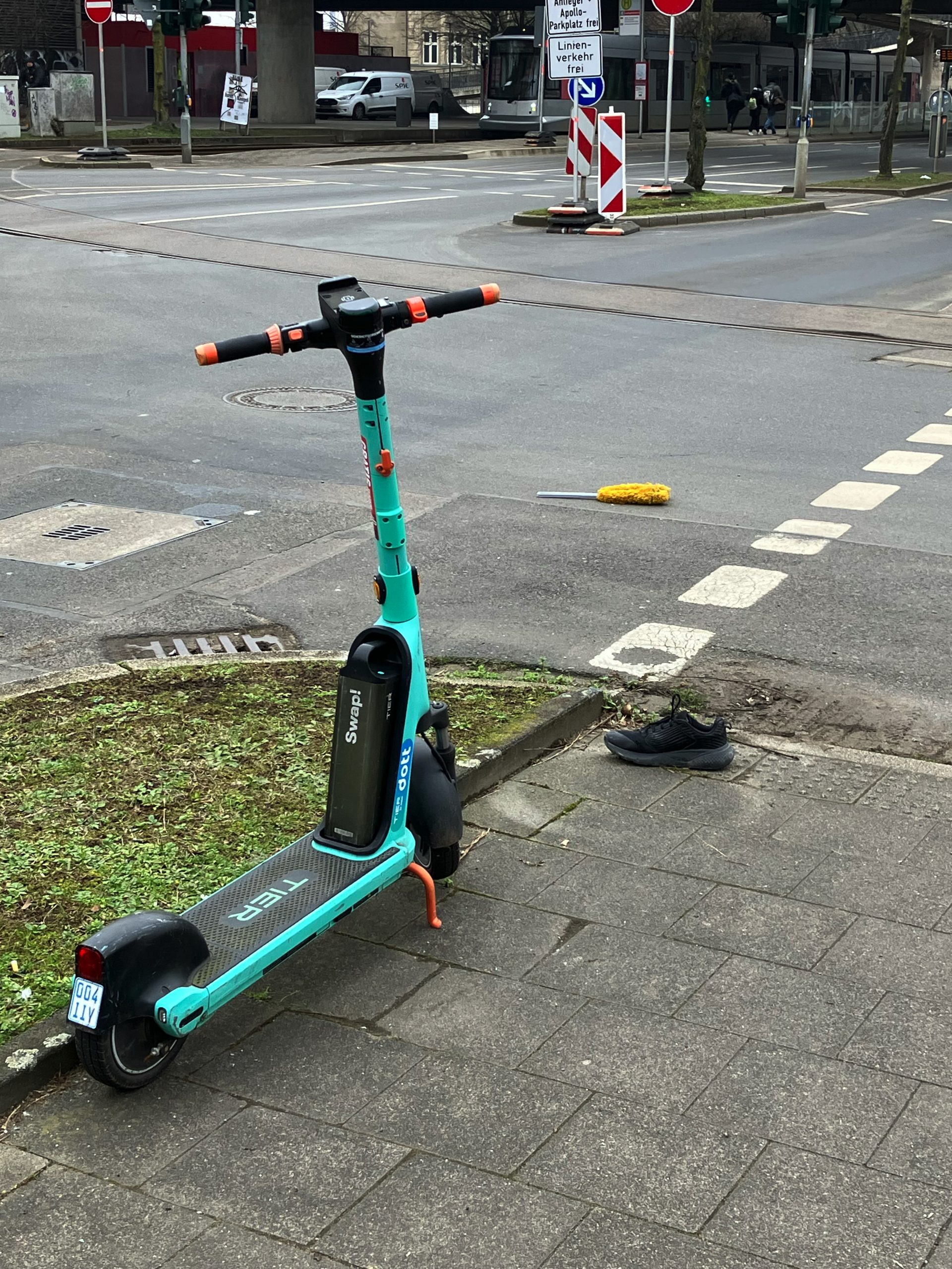 Ein Elektroroller an einer Kreuzung. Davor liegt ein Schuh. Auf der Kreuzung liegt ein Staubwedel.