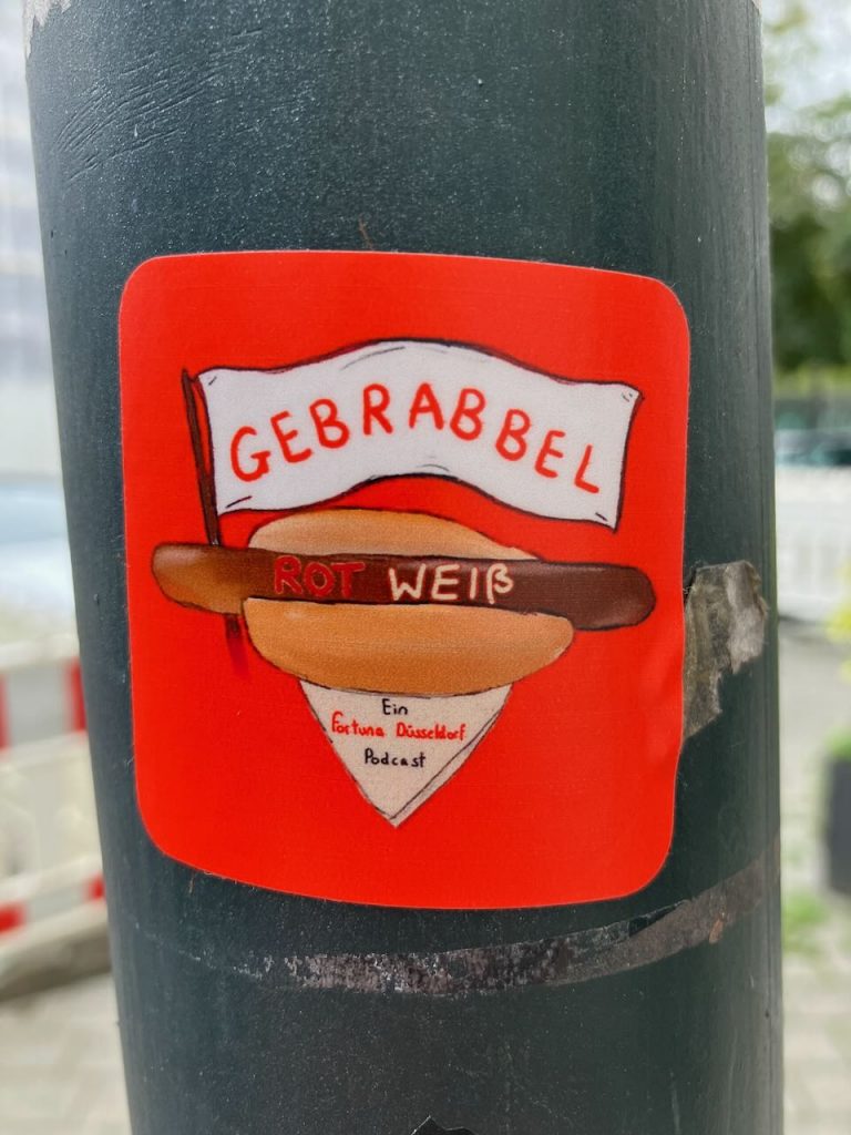 Quadratischer roter Aufkleber. In der Mitte ein gezeichneter Hotdog mit einem weißen Wimpel, darauf steht „GEBRABBEL“. Auf dem Hotdog steht „ROT WEIß“. Unter dem Hotdog ein dreieckiges Papier mit der Aufschrift „Ein Fortuna Düsseldorf Podcast“.