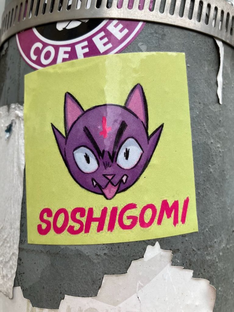 Titel: Lila Katzenkopf mit Schriftzug
Bildbeschreibung: Auf einem gelben Sticker ist ein gezeichneter, lila Katzenkopf mit großen, weißen Augen, spitzen Ohren und herausgestreckter Zunge abgebildet. Über dem Kopf ist ein rotes Kreuz. Darunter steht in pinker, dicker Schrift: “SOSHIGOMI”. Der Sticker klebt auf einer grauen, metallischen Oberfläche.