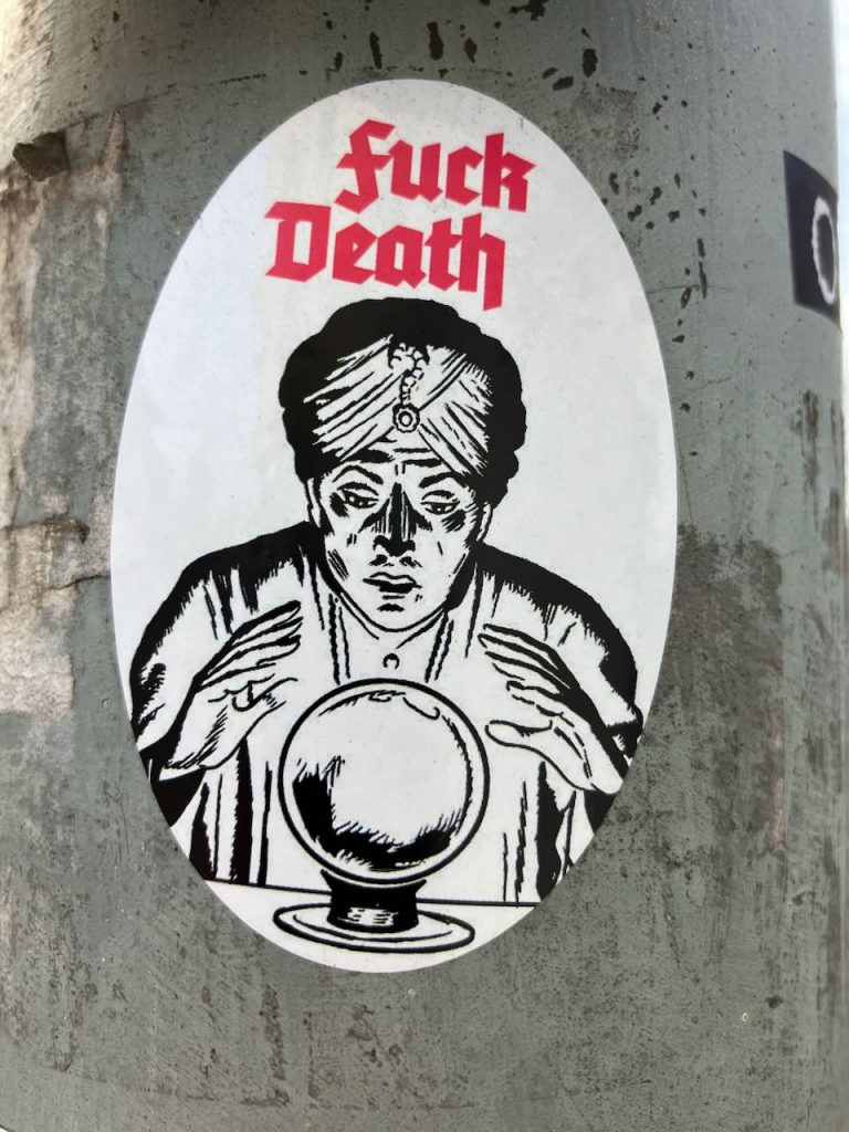 Auf einem Aufkleber ist eine Wahrsagerin mit Turban zu sehen, die ihre Hände über eine große Kristallkugel hält. Darüber steht in roten, auffälligen Buchstaben der Schriftzug „fuck Death“. Der Hintergrund ist schlicht weiß, der Aufkleber befindet sich auf einer grauen, rauen Oberfläche.