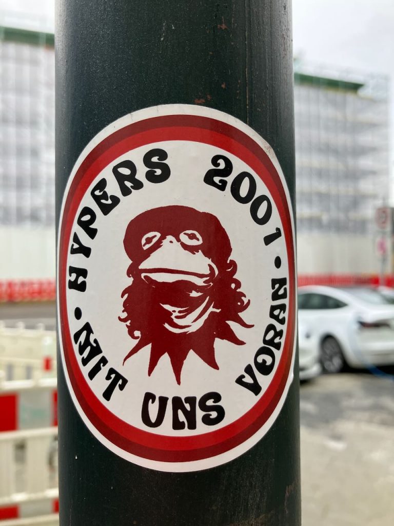 Runder Aufkleber mit rotem Rand. In der Mitte eine stilisierte, dunkelrote Zeichnung eines Froschkopfs mit Mütze und langen Haaren. Im weißen Außenring steht in schwarzer Schrift: „HYPERS 2001 -  MIT UNS VORAN - “.