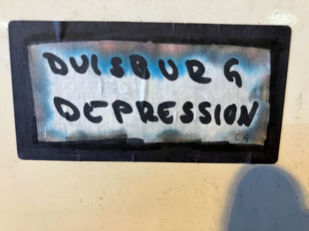 Bildbeschreibung: Ein rechteckiger Aufkleber mit schwarzem Rand auf hellem Untergrund. In der Mitte steht in schwarzen, handgeschriebenen Großbuchstaben „DUISBURG DEPRESSION“. Der Hintergrund des Textes ist silbrig mit blauen und rötlichen Farbschatten. Unten rechts ist ein kleines „C4“ zu sehen.