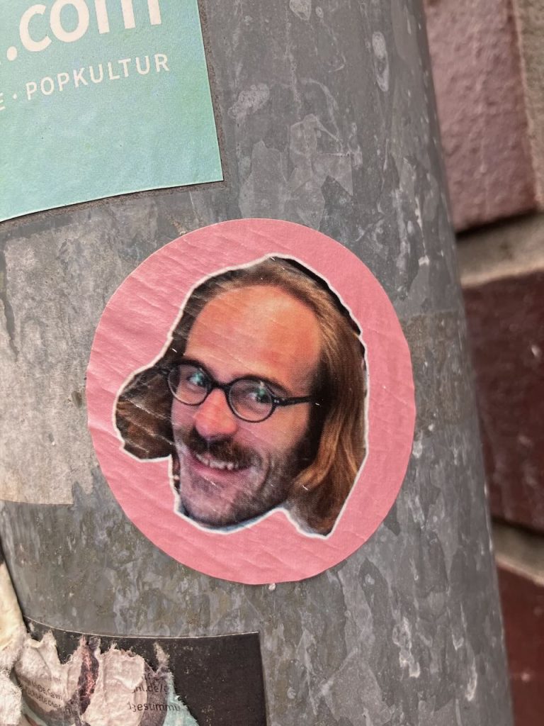 Titel: Sticker mit rosa Rand auf grauem Mast
Bildbeschreibung: Auf einem grauen Metallmast klebt ein runder Sticker mit rosa Rand. In der Mitte ist ein Teil eines Fotos eines Mann mit langen Haaren und Brille mit braunen und beigen Farbtönen zu sehen. Oben links befindet sich ein weiteres, eckiges, türkisfarbenes Stickerfragment mit weißer Schrift. Rechts im Hintergrund sind rote Backsteine sichtbar.