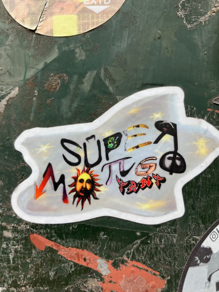 Sticker auf grünem, abgenutztem Untergrund. Der Sticker ist weiß umrandet und zeigt bunte, stilisierte Buchstaben und Symbole, darunter das Wort „SUPER“ in Schwarz, ein rotes Zickzack, eine Sonne, das Pi-Zeichen, eine geballte Faust und das Wort „PANT“ in roter Schrift. Im Hintergrund sind gelbe Sterne verteilt.
Titel: Bunter SUPER-Sticker mit Symbolen
