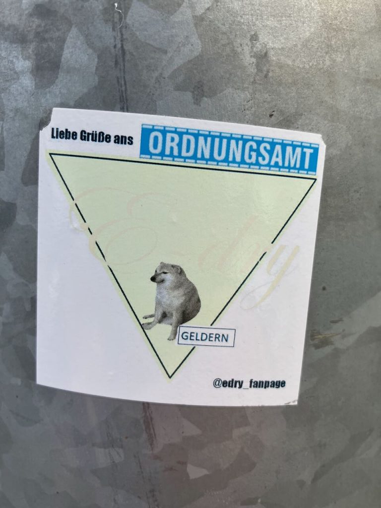 Sticker auf grauem Untergrund. Oben steht „Liebe Grüße ans ORDNUNGSAMT“. In der Mitte ein auf dem Boden sitzender Hund in einem nach unten zeigenden Dreieck. Unter dem Hund steht „GELDERN“. Unten rechts ist der Instagram-Name „@edry_fanpage“.