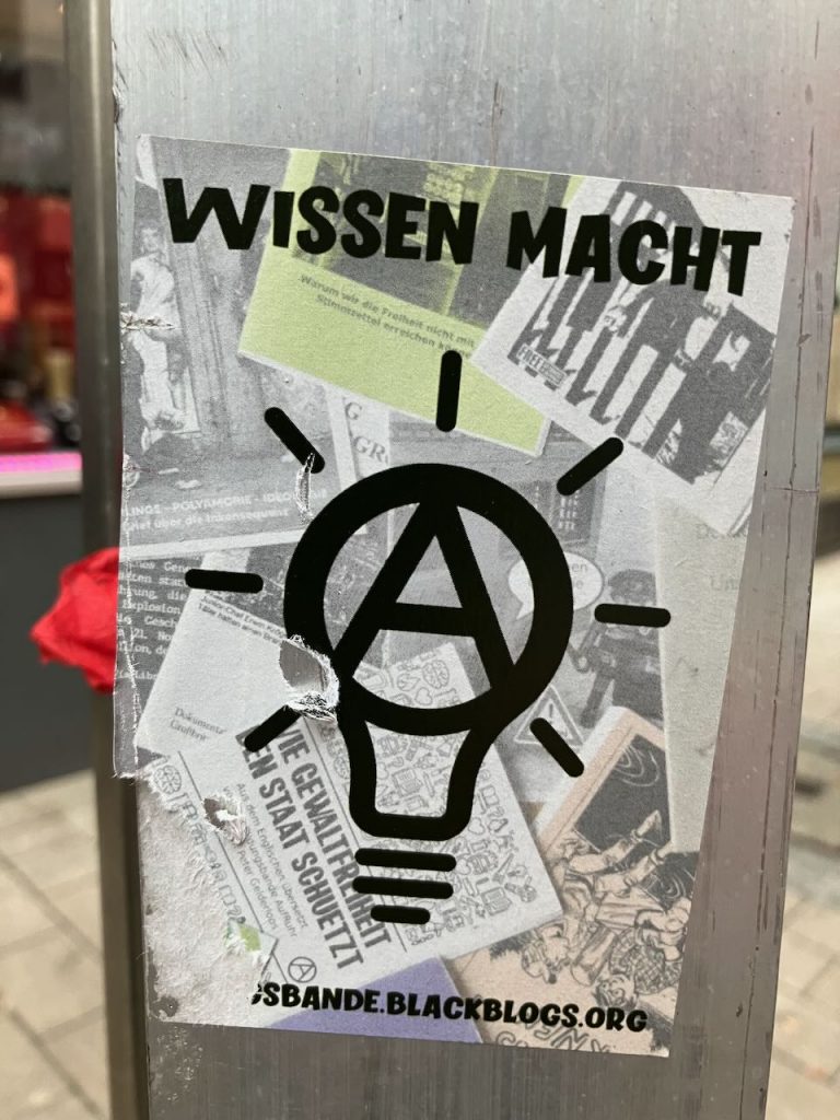 Titel: Wissen als Macht
Bildbeschreibung: Ein Aufkleber auf Metall zeigt den Text „WISSEN MACHT“ und eine stilisierte Glühbirne mit Anarchie-A im Zentrum. Im Hintergrund sind schwarz-weiße Collagen aus Zeitungsartikeln und Zeichnungen zu sehen. Unten steht: „SBANDE.BLACKBLOGS.ORG“.