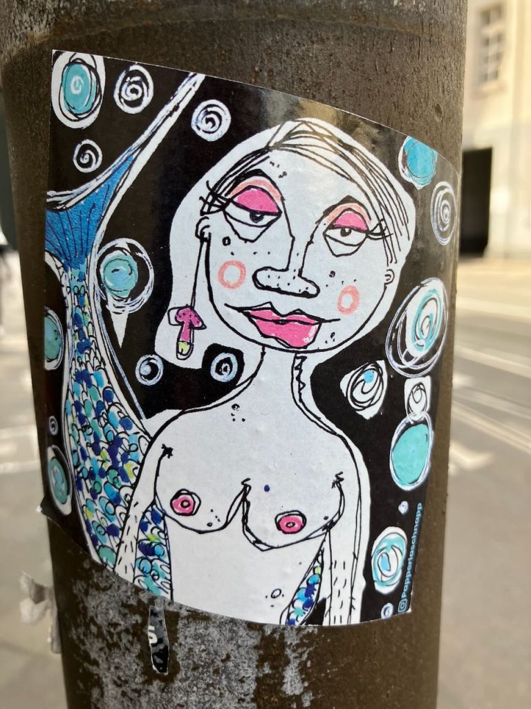 Bunter Aufkleber an einem Mast. Abgebildet ist eine gezeichnete Meerjungfrau mit nacktem Oberkörper, rosa Brustwarzen und blau-grün schuppigem Fischschwanz. Im Hintergrund sind viele weiße und blaue Kreise auf schwarzem Grund. In der unteren rechten Ecke steht ein Instagram-Name.