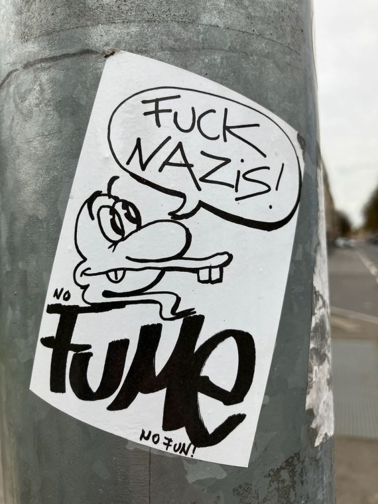 Titel: Sticker mit Comicfigur und Schriftzug

Bildbeschreibung: Ein schwarz-weißer Sticker klebt an einem grauen Laternenpfahl. Darauf ist eine Comicfigur mit großer Nase zu sehen, die mit dem Finger zeigt. In einer Sprechblase steht „FUCK NAZIS!“. Darunter steht groß das Wort „FUME“, daneben „NO FUN!“.