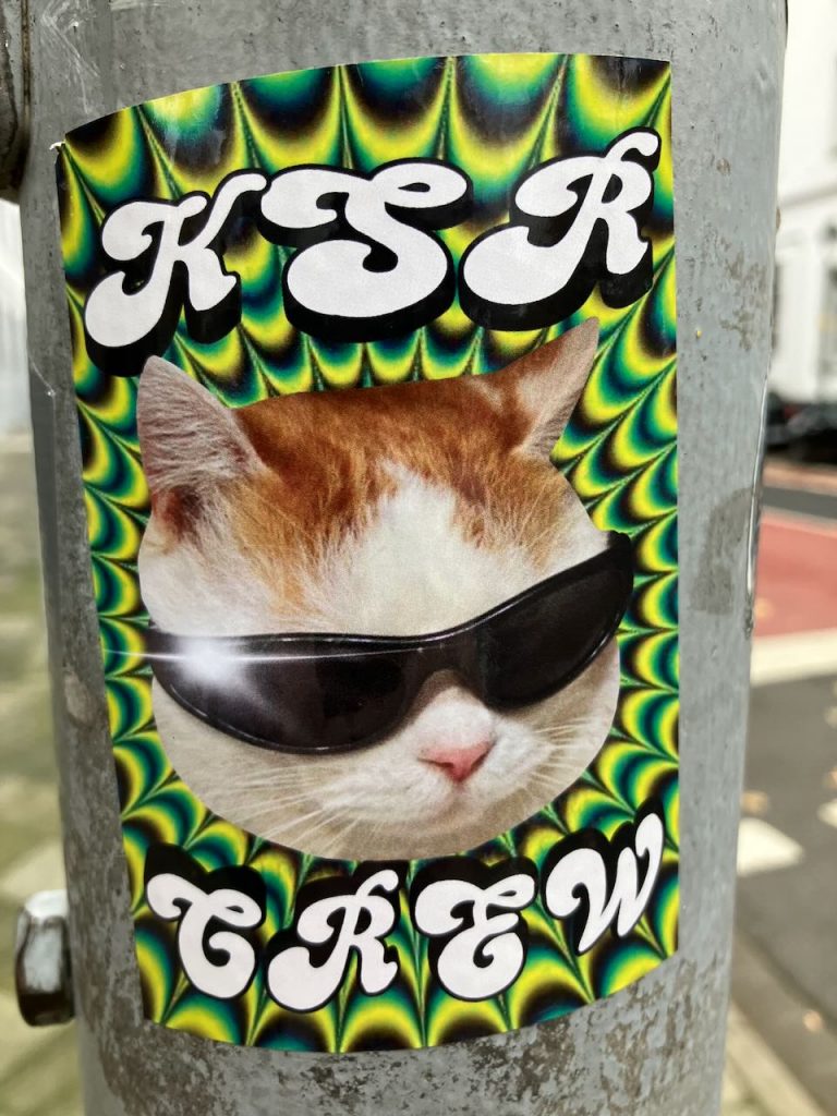 Titel: Katze mit Sonnenbrille – KSR CREW
Bildbeschreibung: Auf einem Sticker ist eine rot-weiße Katze mit schwarzer Sonnenbrille zu sehen. Der Hintergrund ist bunt und psychedelisch gemustert. Oben steht in großen weißen Buchstaben „KSR“, unten „CREW“. Der Sticker klebt an einem grauen Metallpfosten.