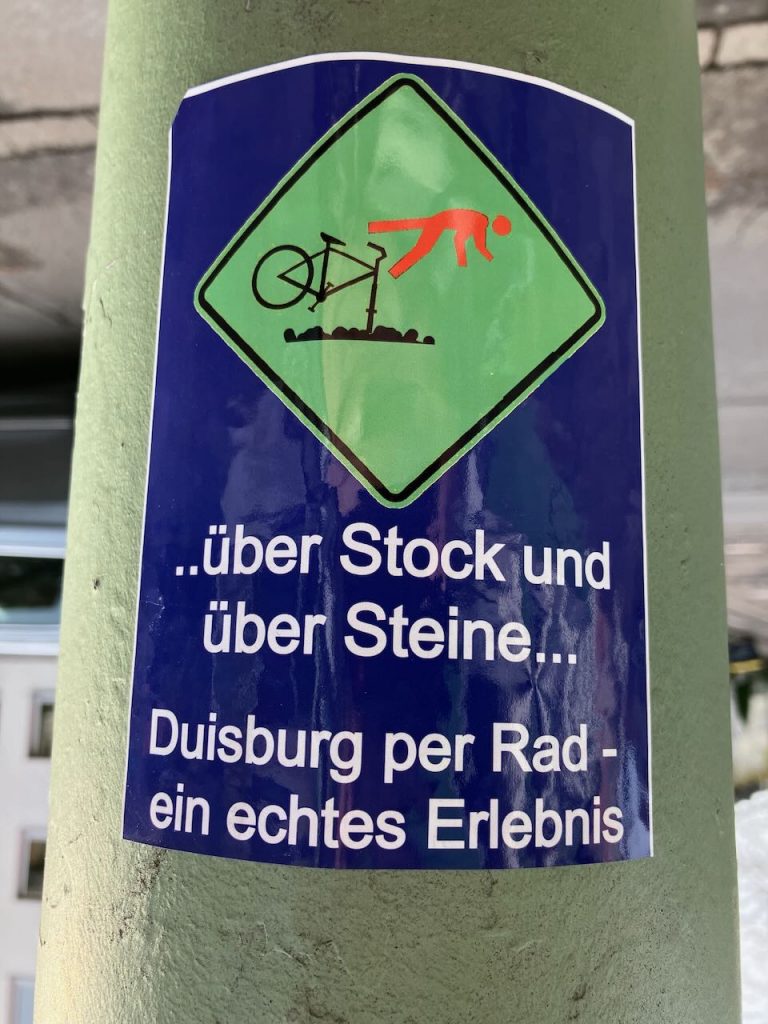 Auf einem blauen Aufkleber ist ein grünes Verkehrsschild abgebildet. Darauf stürzt eine stilisierte Person in Rot über den Lenker eines Fahrrads, das über Steine fährt. Darunter steht in weißer Schrift: „..über Stock und über Steine… Duisburg per Rad – ein echtes Erlebnis“.