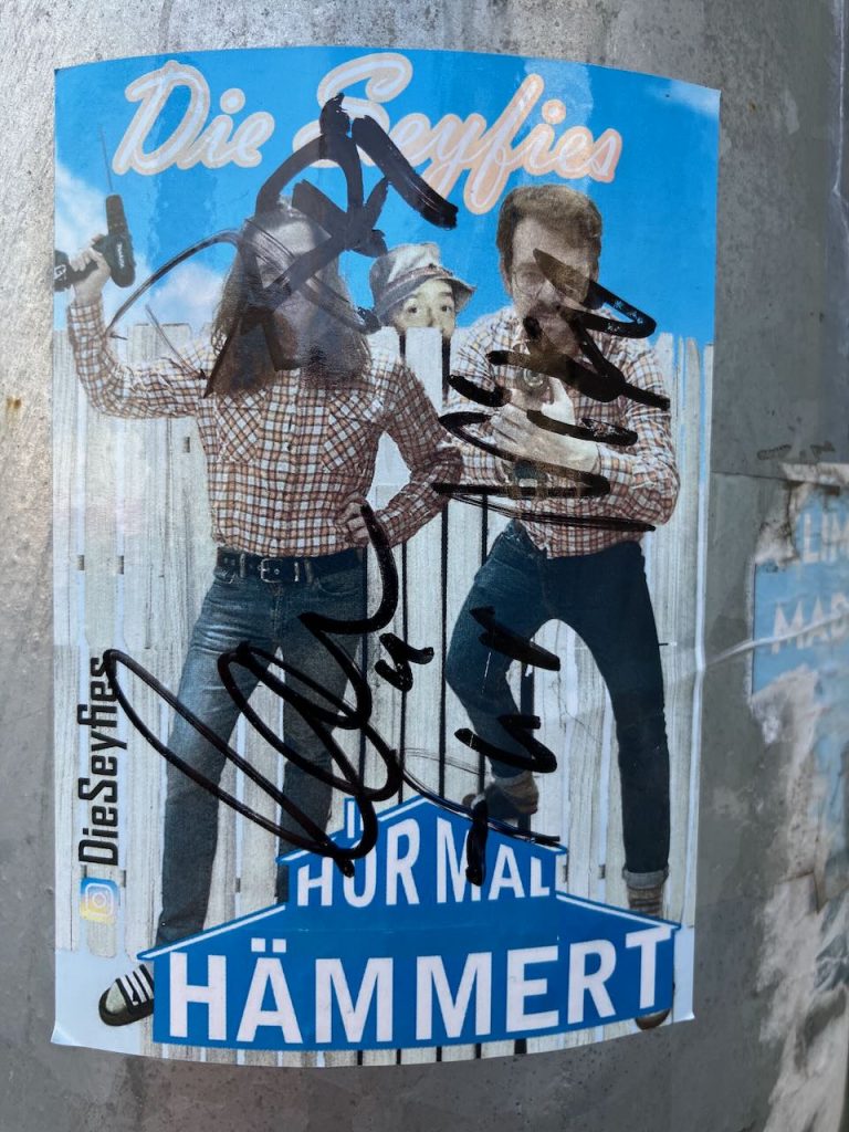 Aufkleber mit zwei Personen in karierten Hemden und Jeans, eine hält einen Hammer. Im Hintergrund ein weißer Zaun, oben steht in gelber Schrift „Die Seyfies“. Unten auf einem blauen Schild in weißer Schrift: „HÖR MAL HÄMMERT“. Schwarze Schriftzüge und Unterschriften sind über das Bild gemalt. Links am Rand steht „DieSeyfies“ mit Instagram-Logo.