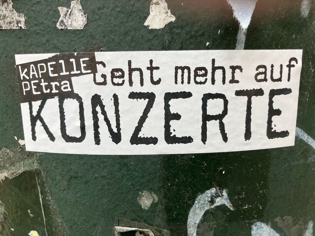 Weißes rechteckiges Sticker auf grünem, abgenutztem Untergrund. Darauf steht in schwarzer, großer und kleiner Schrift: “KAPELLE PETRA Geht mehr auf KONZERTE”.