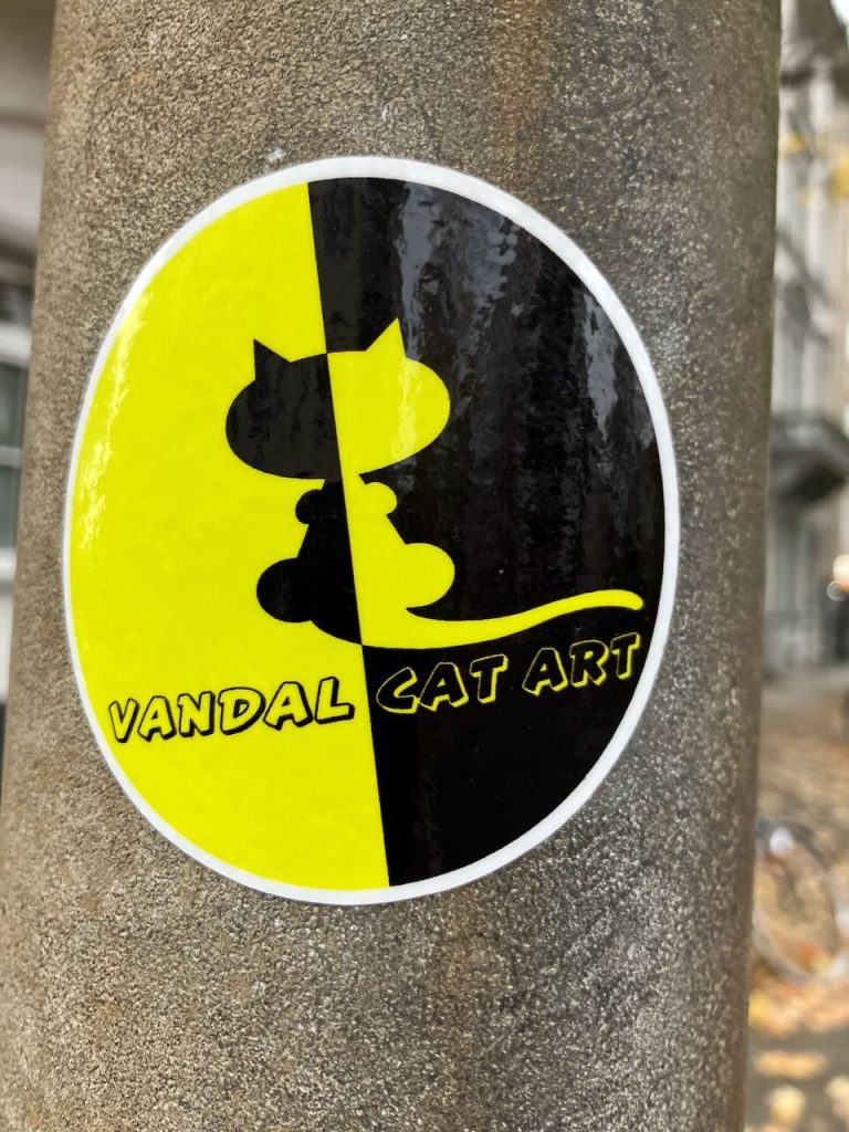 Titel: Sticker „Vandal Cat Art“
Bildbeschreibung: Ein runder Aufkleber auf einem grauen Mast. Der Sticker ist gelb und schwarz halbiert. In der Mitte ist eine stilisierte Katze, deren Umriss sich farblich absetzt. Unten steht in gelber und schwarzer Schrift: „VANDAL CAT ART“.