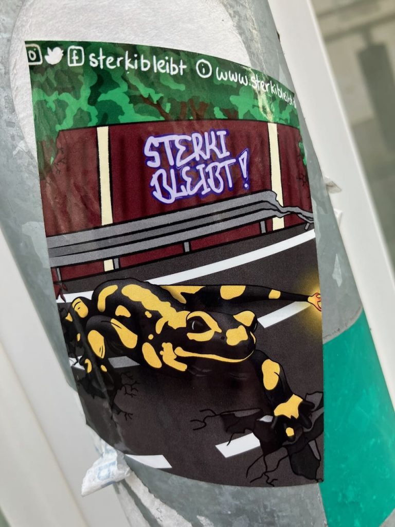 Auf einem Aufkleber ist ein schwarz-gelb gefleckter Salamander auf einer Straße abgebildet. Im Hintergrund sind grüne Bäume und ein rotes Gebäude zu sehen. Über dem Gebäude steht in blauer Schrift „STERKI BLEIBT!“. Am oberen Rand sind Social-Media-Symbole und die Webadresse „sterkibleibt“.
