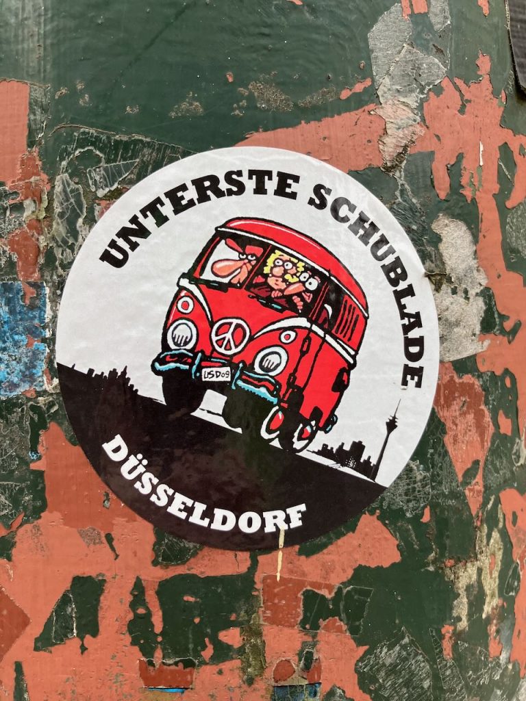 Runder Aufkleber auf grün-orange-braunem Untergrund. In der Mitte ein roter VW-Bus mit mehreren gezeichneten Personen, die aus dem Fenster schauen. Auf dem Bus steht „LSD 09“. Im Hintergrund eine schwarze Stadtsilhouette mit Fernsehturm. Oben steht in schwarzer Schrift „UNTERSTE SCHUBLADE“, unten „DÜSSELDORF“.