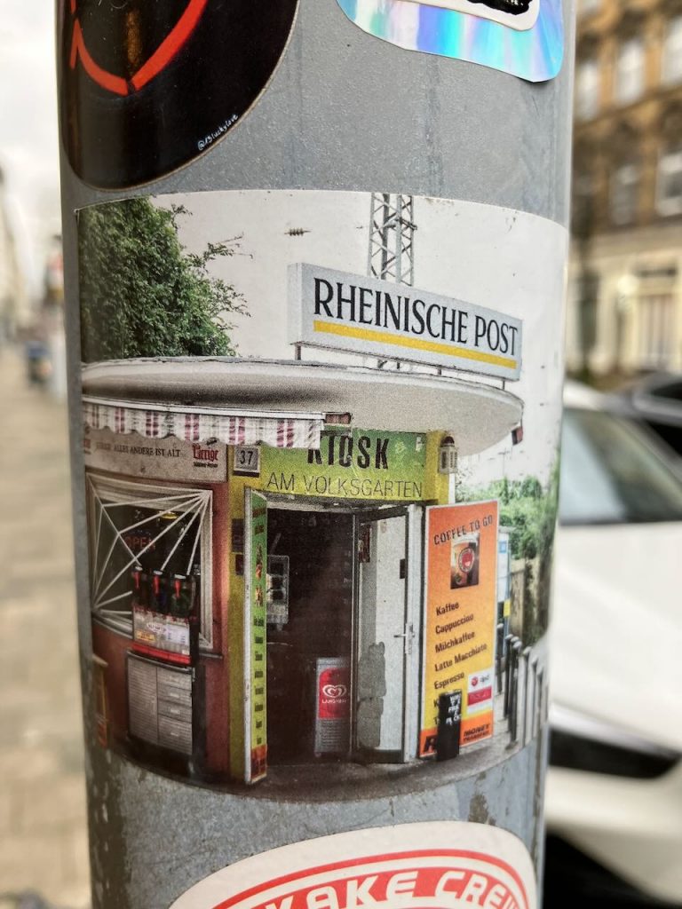 Titel: Kiosk am Volksgarten
Bildbeschreibung: Auf einem grauen Laternenpfahl klebt ein Aufkleber mit dem Foto eines kleinen Kiosks. Über dem runden Dach steht ein großes Schild „RHEINISCHE POST“. Am Eingang ist ein grünes Schild mit „Kiosk am Volksgarten“. Rechts hängt ein oranges Schild mit Kaffeeangeboten. Vor dem Kiosk stehen Automaten und ein Eisschild.