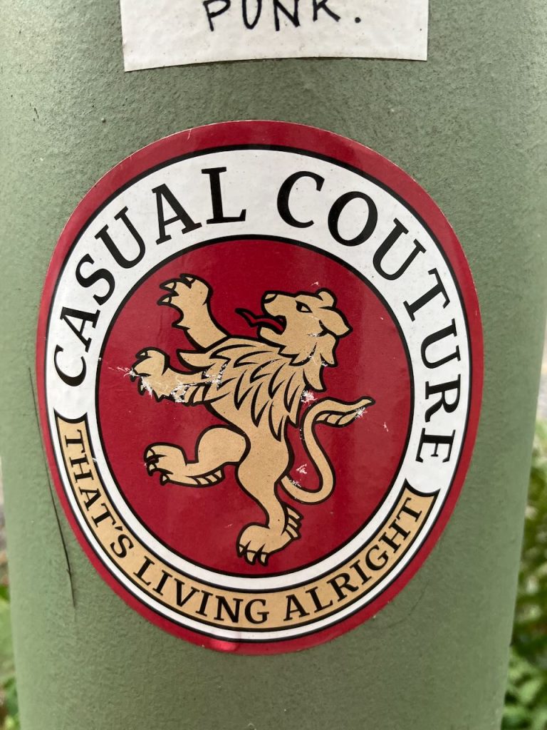 Runder Aufkleber auf grünem Untergrund. In der Mitte ein goldener, aufrecht stehender Löwe auf rotem Hintergrund. Außen ein weißer Ring mit schwarzer Schrift: Oben steht „CASUAL COUTURE“, unten „THAT’S LIVING ALRIGHT“. Am oberen Rand des Bildes ein kleiner weißer Aufkleber mit handgeschriebenem Text „PUNK.“
