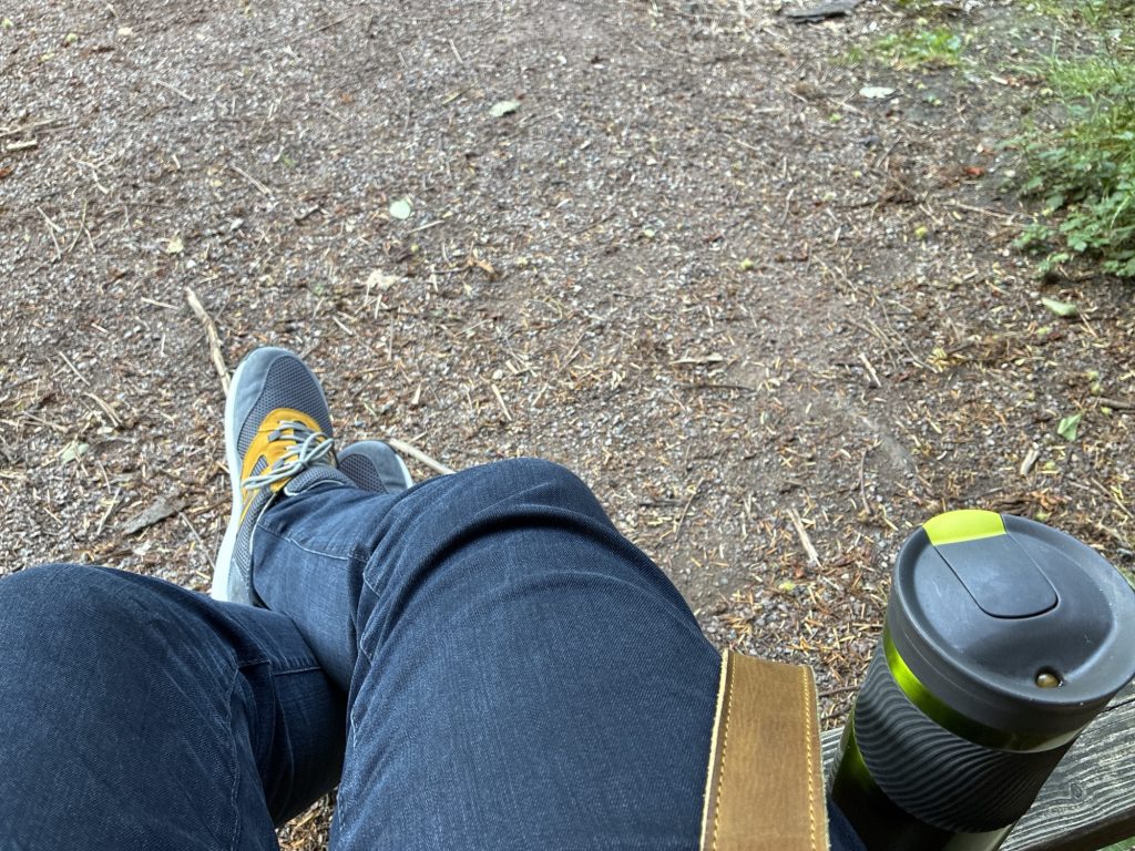 Waldboden, Beine in Jeanshose, Schuhe und ein Thermobecher auf einer Sitzbank.
