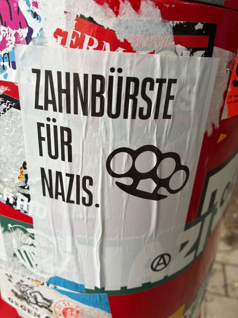 Weißer Sticker mit schwarzer Schrift: „ZAHNBÜRSTE FÜR NAZIS.“ Daneben ist eine schwarze Illustration eines Schlagrings zu sehen. Unten rechts ein kleines Anarchie-Symbol.