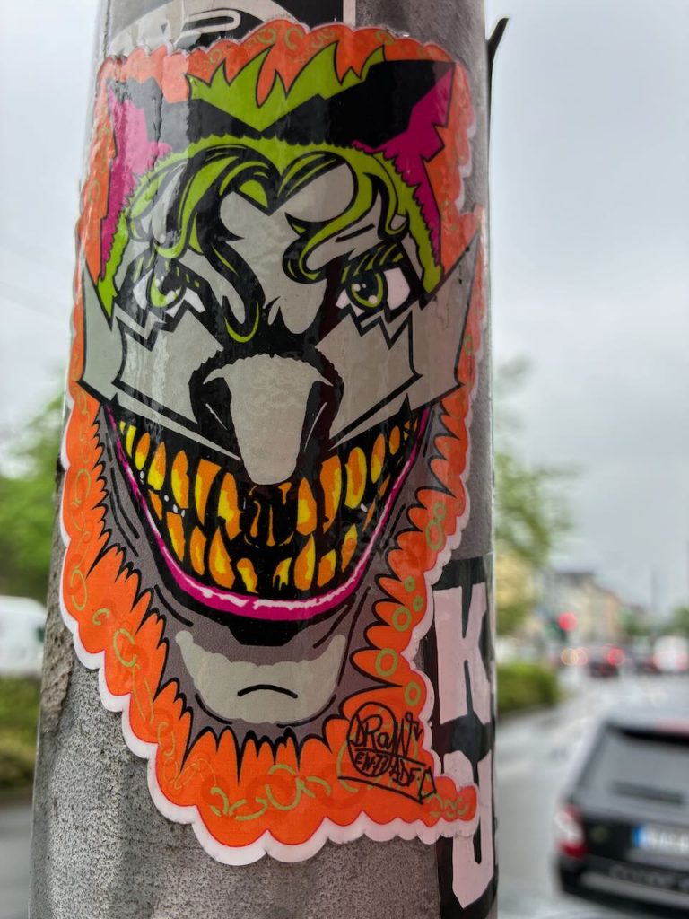Bunter Sticker mit einem stilisierten, grinsenden Gesicht, das an den Joker erinnert. Die Zähne sind groß und gelb, das Gesicht ist von kräftigen Farben wie Grün, Orange und Pink umrahmt.