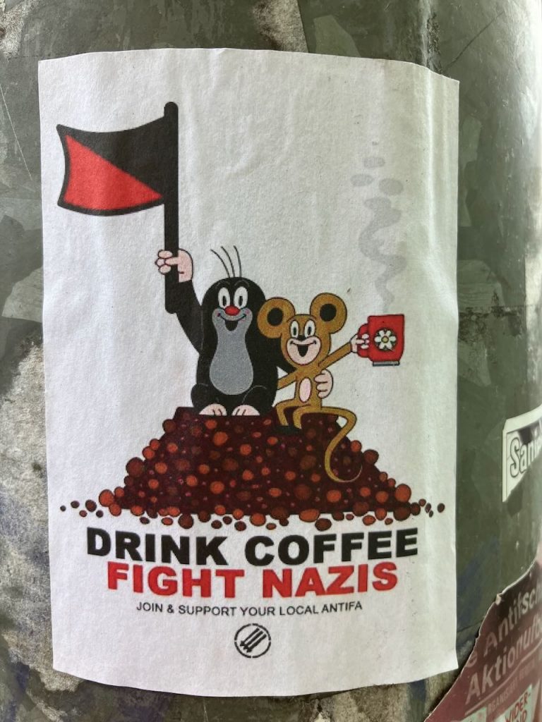 Zwei Comicfiguren, ein Maulwurf und eine Maus, sitzen auf einem Haufen Kaffeebohnen. Der Maulwurf hält eine schwarz-rote Fahne, die Maus eine Kaffeetasse. Darunter steht: „DRINK COFFEE FIGHT NAZIS – JOIN & SUPPORT YOUR LOCAL ANTIFA“.
