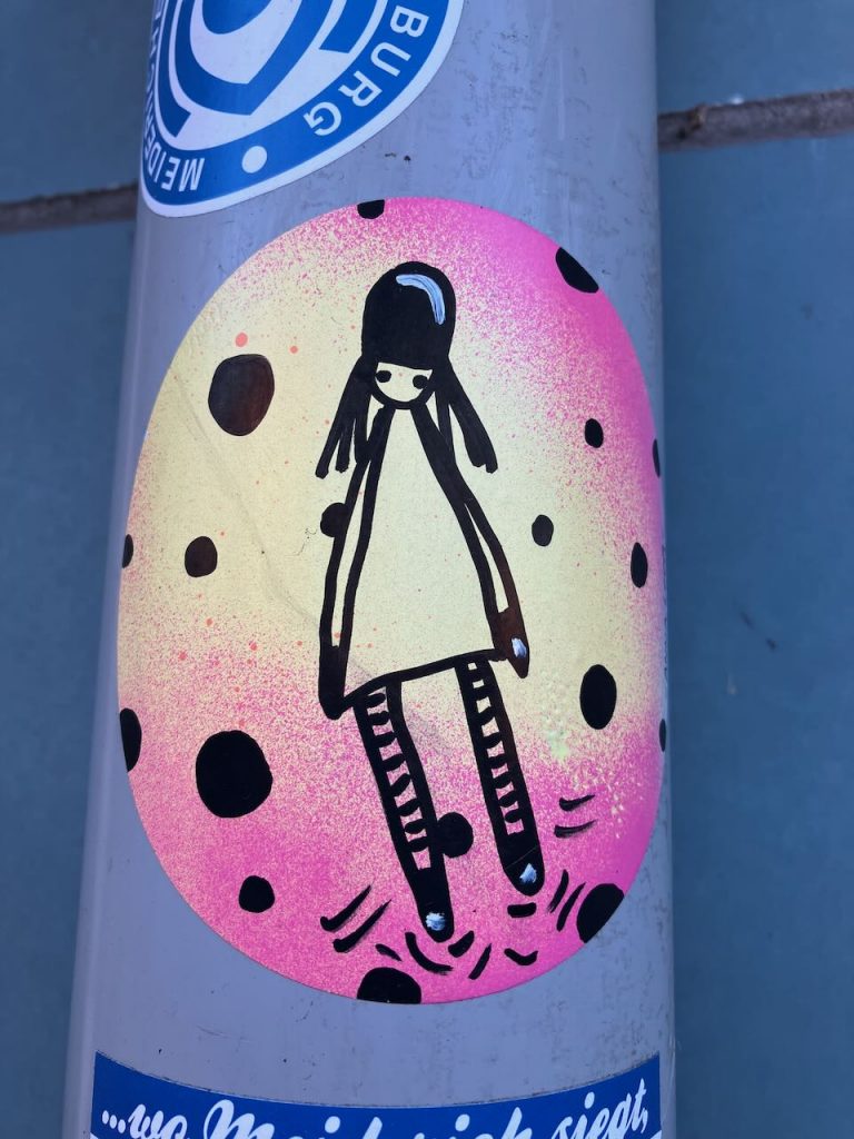 Ein Aufkleber zeigt eine stilisierte Figur eines Mädchens mit langen Haaren und gestreiften Strümpfen. Der Hintergrund ist rund, gelb-pink verlaufend mit schwarzen Punkten.