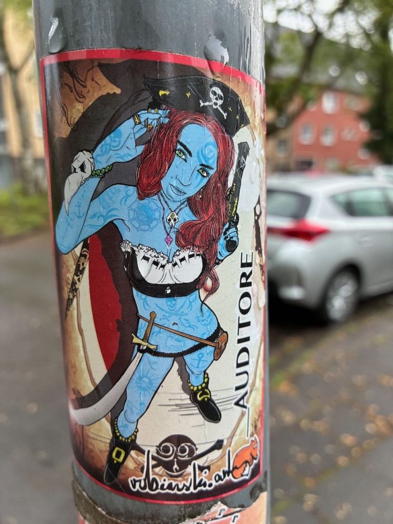 Farbiger Sticker einer tätowierten Piratin mit rotem Haar, Piratenhut, Säbel und Pistole. Im Hintergrund ist ein Totenkopf zu sehen, seitlich steht „AUDITORE“ und unten eine Signatur.