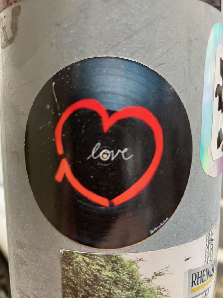Ein runder Sticker auf grauem Untergrund zeigt eine schwarze Schallplatte mit einem roten, handgezeichneten Herz. In der Mitte steht in weißer Schrift das Wort „love“. Das Motiv wirkt schlicht und freundlich.