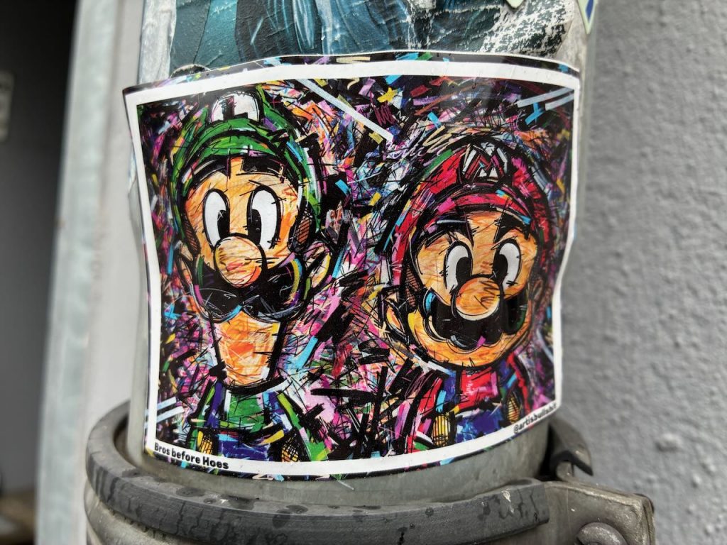 Bunt und wild gezeichneter Sticker mit den Figuren Luigi (links, grün) und Mario (rechts, rot) aus der bekannten Videospielreihe. Beide haben große Augen, schwarze Schnurrbärte und stehen nebeneinander vor einem abstrakten, farbenfrohen Hintergrund. Unten steht: “Bros before Hoes”.