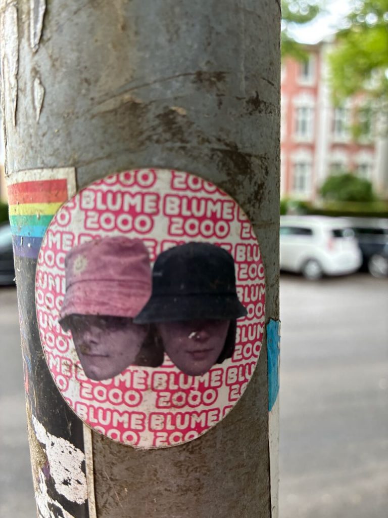 Runder Sticker mit weiß-rotem Hintergrund und dem wiederholten Text „BLUME 2000“. Im Vordergrund sind zwei Personen mit Kopfbedeckungen zu sehen.