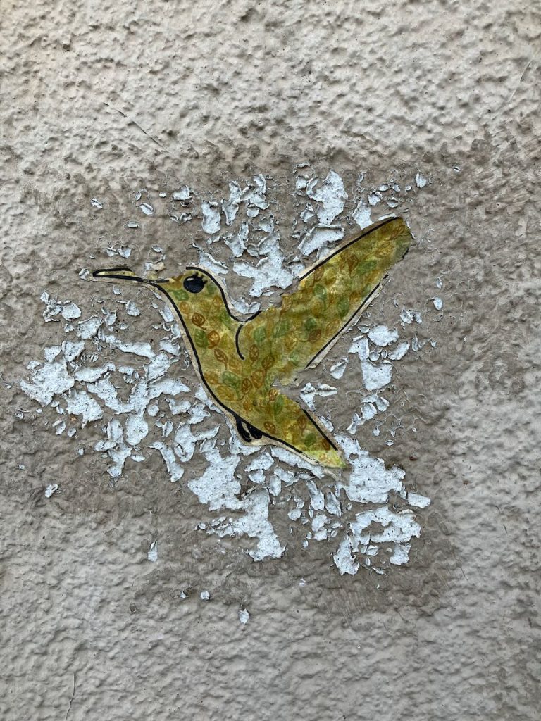 Ein kleiner, gelber Kolibri ist auf eine raue, abgeplatzte Wand gemalt. Der Vogel ist seitlich dargestellt, der Hintergrund ist weiß und bröckelig.