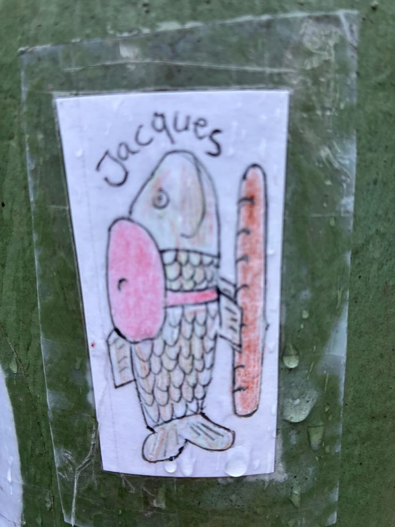 Handgezeichneter Sticker eines Fisches mit rosa Kiemen und Halsband, daneben ein Baguette. Darüber steht „Jacques“. Der Hintergrund ist weiß, der Sticker ist mit Klebeband befestigt.