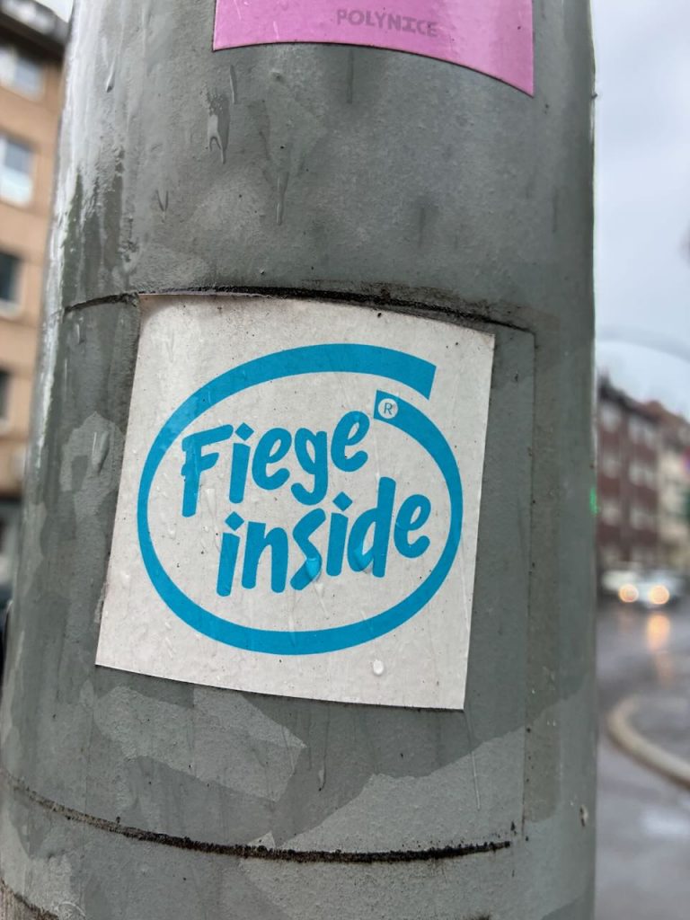 Weißer Sticker mit blauem Schriftzug „Fiege inside“ in einem ovalen Rahmen, angelehnt an das bekannte „Intel inside“-Logo.