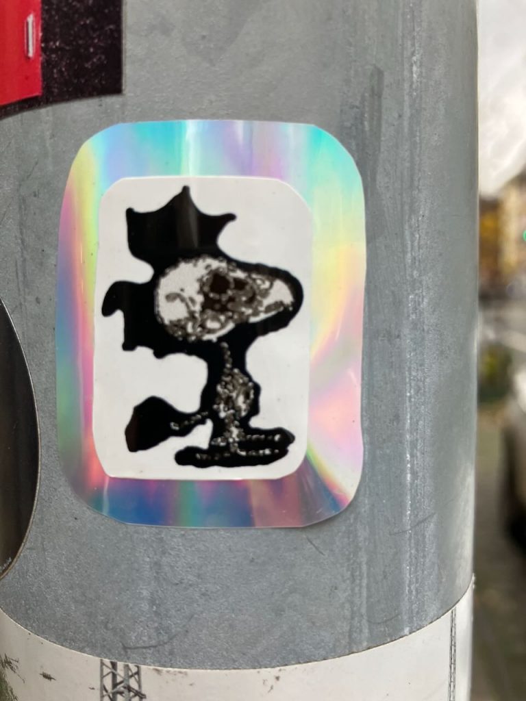 Ein eckiger Sticker mit holografischem Rand zeigt den Vogel Woodstock als Röntgenbild.