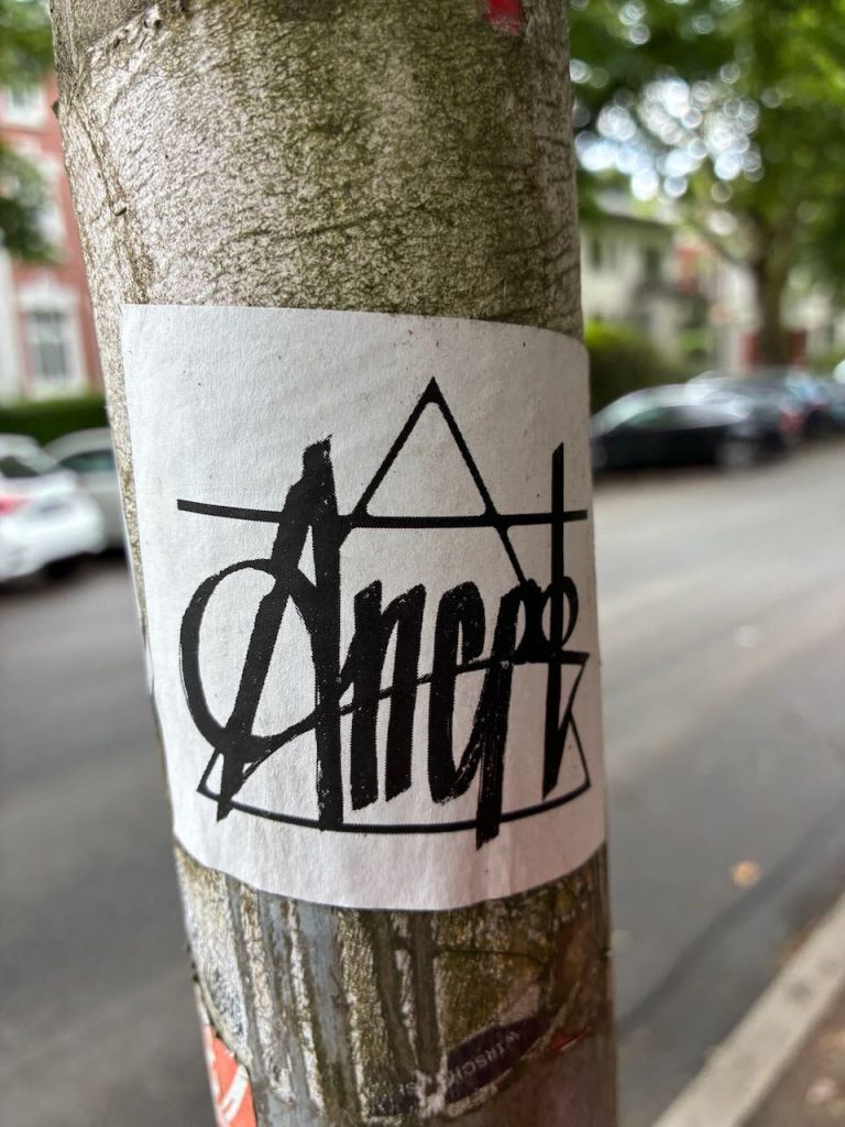 Weißer Sticker mit schwarzem, grafisch gestaltetem Schriftzug, der das Wort „Ancap“ in einer Mischung aus Druck- und Schreibschrift darstellt. Im Hintergrund geometrische Linien.