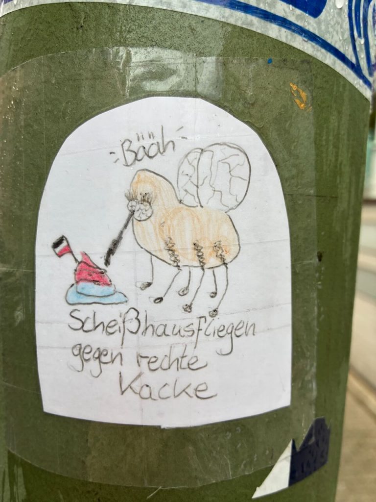 Handgezeichneter Sticker mit einer Fliege, die vor einem Haufen mit Reichsfahne steht. Über ihr steht: "Bääh!" Text: „Scheißhausfliegen gegen rechte Kacke“.