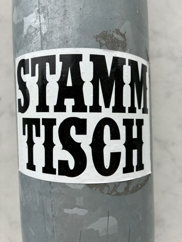 Weißer Sticker mit dem Wort „STAMMTISCH“ in fetten, schwarzen Großbuchstaben, mittig auf einem grauen Pfosten angebracht.
