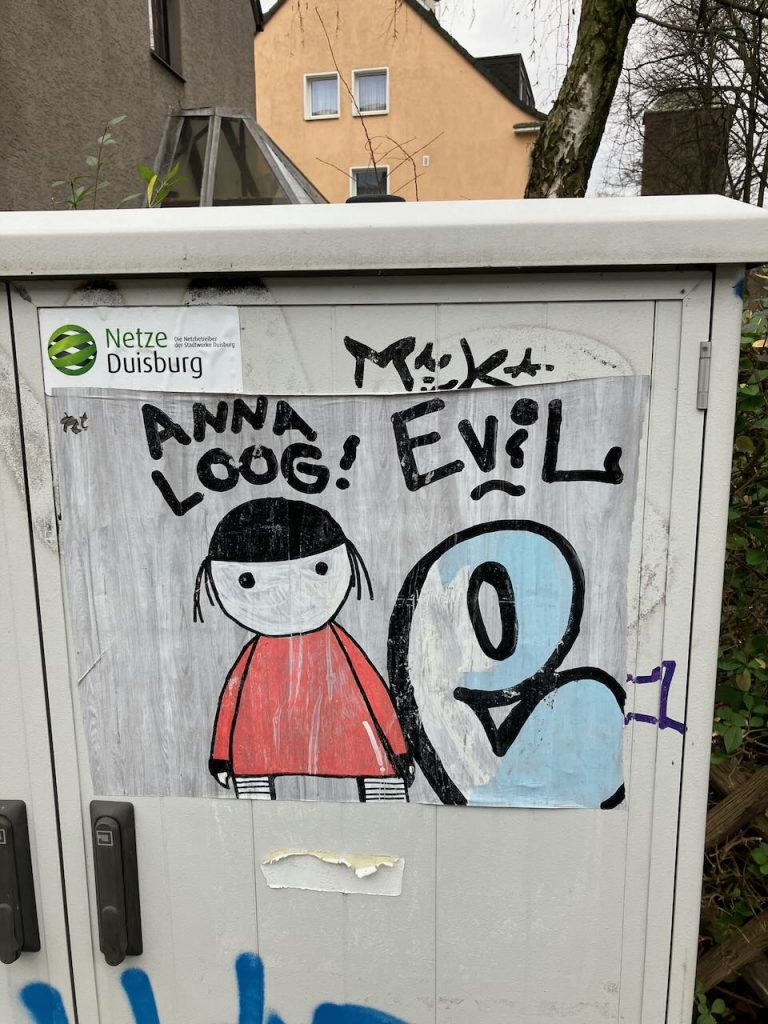 Eine einfache Zeichnung zeigt ein Mädchen mit schwarzen Haaren und rotem Kleid neben einer großen, blauen, stilisierten Figur mit Gesicht. Darüber steht: „ANNA LOOG! EVIL“.