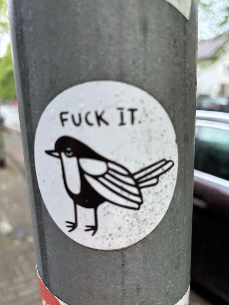 Runder, schwarz-weißer Sticker mit einer stilisierten Elster. Über dem Vogel steht in Großbuchstaben „FUCK IT.“.