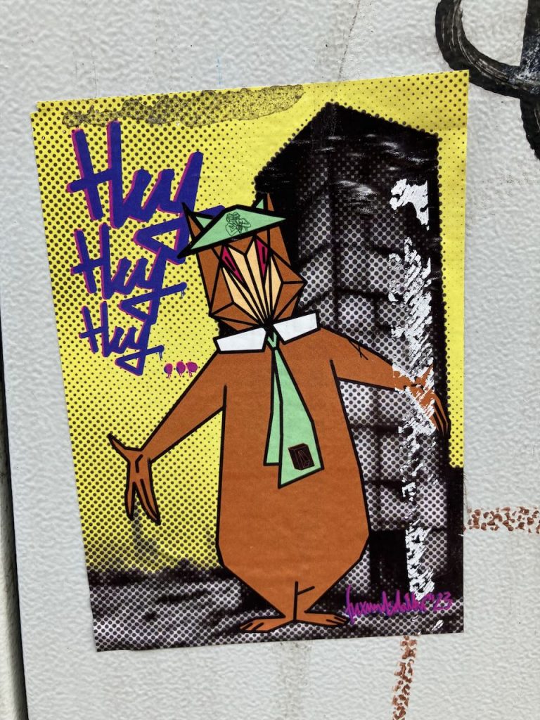 Der Sticker zeigt Yogi Bär, jedoch stark verfremdet: Die Figur ist vereinfacht und im Cartoon-Stil gehalten, mit braunem Fell, grünem Hut und grüner Krawatte. Die Gesichtszüge sind reduziert, die Formen kantiger und abstrakter als im Original. Im Hintergrund steht mehrfach in lila Schrift das Wort „Hey“. Die Verfremdung betont klare Flächen, kräftige Farben und eine poppige, moderne Wirkung.