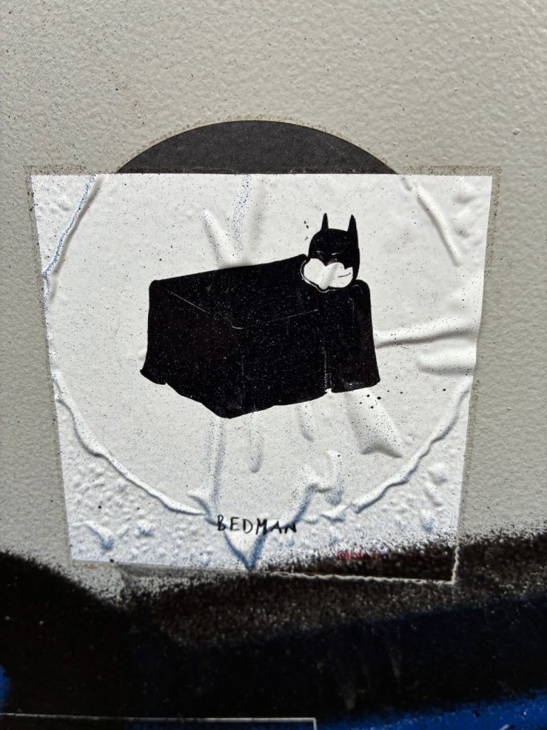 Weißer Sticker mit einer schwarzen Illustration: Ein Bett mit Batman-Maske, darunter der Schriftzug „BEDMAN“.