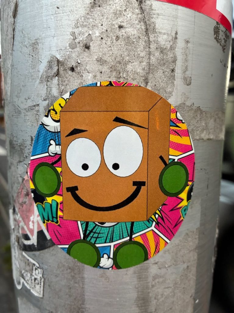 Runder Sticker mit bunter Comic-Grafik: Ein brauner Würfel mit Gesicht, Armen und Beinen, im Hintergrund bunte Pop-Art-Elemente.
