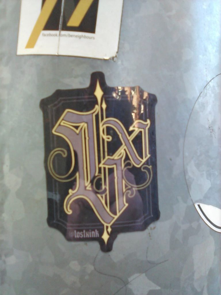 Ein Sticker mit einem goldgelben, verschnörkelten Monogramm auf dunklem Hintergrund. Unten steht klein: „lost1xink“.