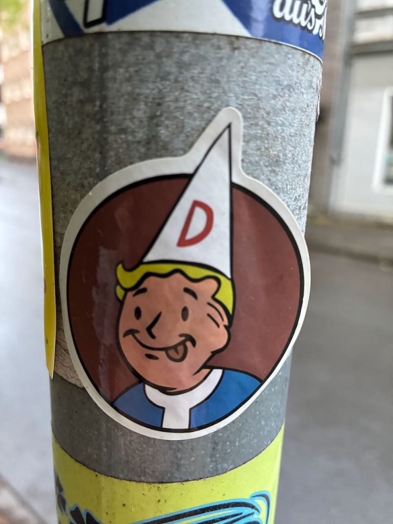 Ein runder Sticker zeigt eine Comicfigur mit blonden Haaren und einer weißen Mütze mit rotem „D“. Die Figur lächelt, der Hintergrund ist braun.