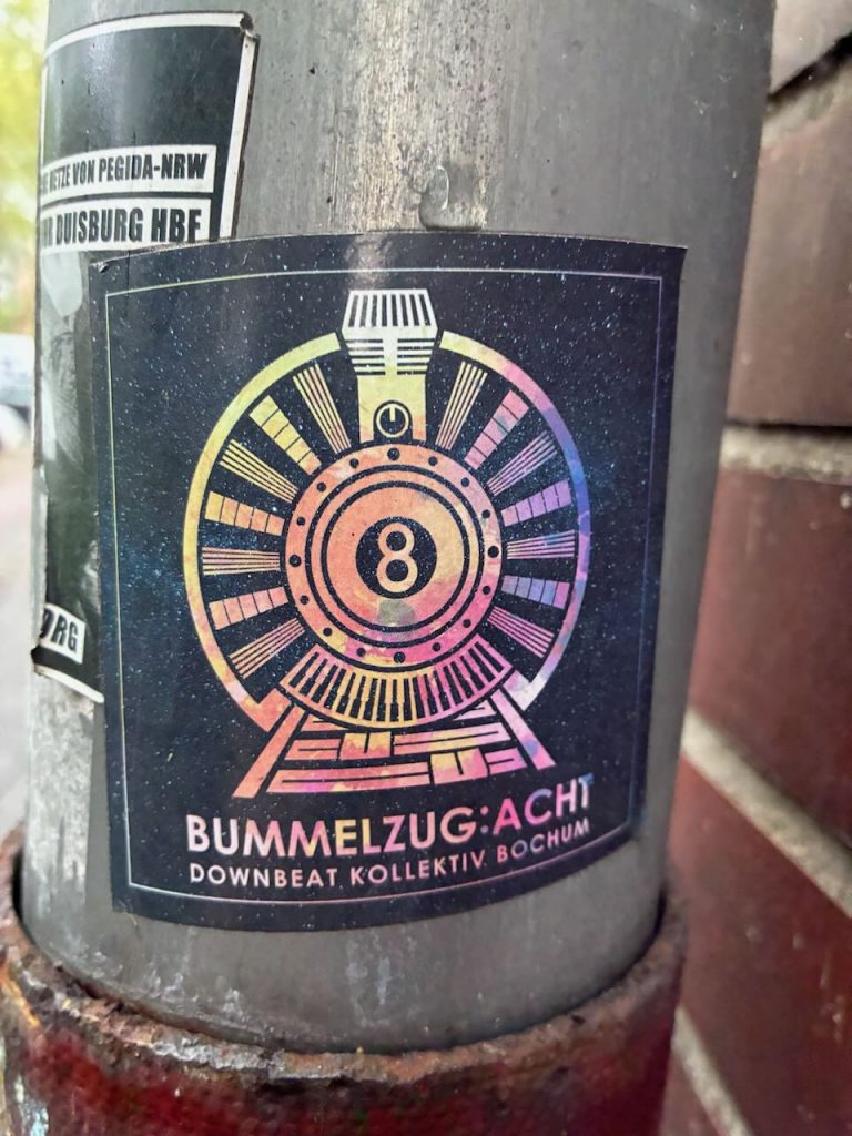 Quadratischer Sticker mit bunter, stilisierter Lokomotive und der Zahl 8 auf der Front. Darunter steht: „BUMMELZUG:ACHT DOWNBEAT KOLLEKTIV, BOCHUM“.