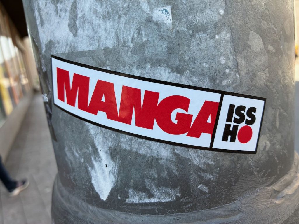 Rechteckiger Sticker im Stil eines Logos. Großes rotes „MANGA“, daneben in Schwarz und Rot „ISSHO“.